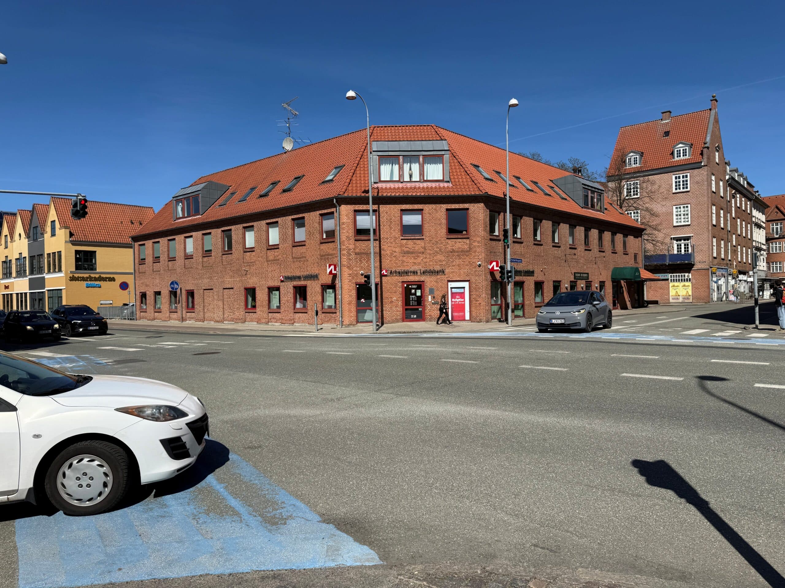 Hjørneejendommen i røde mursten set fra gadehjørnet, med parkeringspladser langs facaden